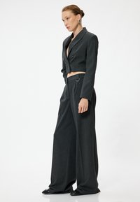 Veste grise cintrée avec des boutons sur le devant, associée à un pantalon gris à jambes larges avec passants de ceinture et une texture subtile. Des chaussures noires complètent le look.