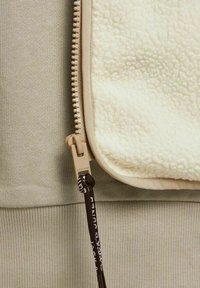 Fermeture éclair beige avec tirette noire attachée à une polaire couleur crème et un tissu côtelé marron clair.