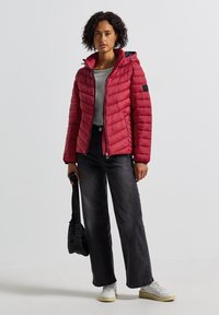 Rode pufferjack met een ritssluiting en een capuchon, voorzien van gestikte details en een klein logopatch, gecombineerd met zwarte wijde jeans.
