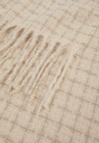 Coperta intrecciata beige con un motivo a griglia testurizzata e bordi frangiati; realizzata in tessuto morbido con un tocco di brillantezza.