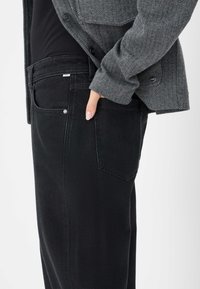Jeans neri con taglio dritto, dotati di una tasca posteriore, dettagli in rivetti metallici e una texture che indica il tessuto denim. Indossati sotto una giacca grigia a motivo zigzag.