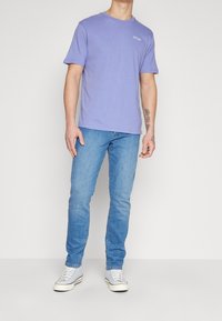 Man som bär en lilarosa kortärmad t-shirt, ljusblå slim-fit jeans, vita sneakers och synliga tatueringar på sin högra underarm och hand.