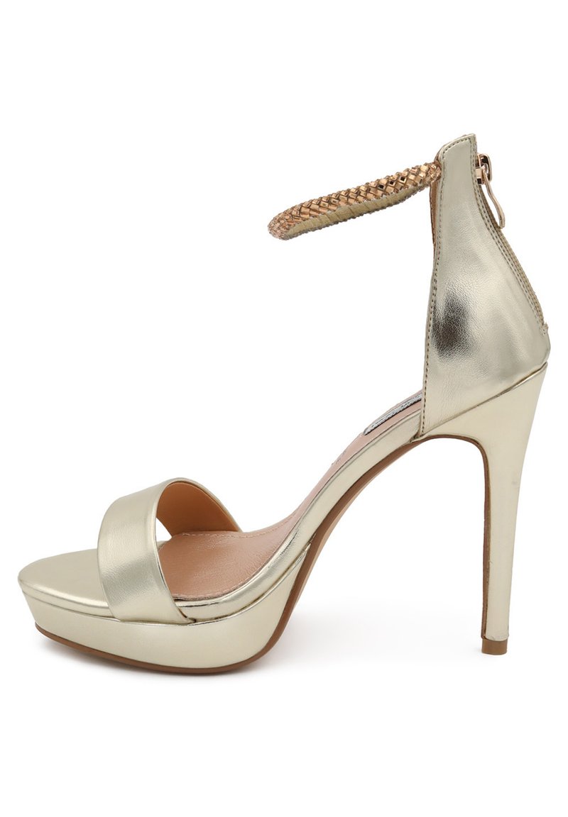 Sandalo con tacco alto in metallo dorato, con punta aperta, ampia fascia frontale e cinturino intrecciato alla caviglia. Presenta una piattaforma per un'altezza aggiuntiva.