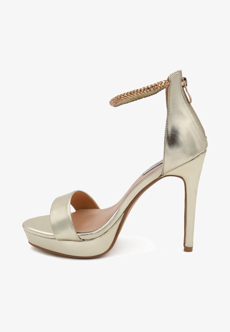 Sandalo con tacco alto in metallo dorato, con punta aperta, ampia fascia frontale e cinturino intrecciato alla caviglia. Presenta una piattaforma per un'altezza aggiuntiva.