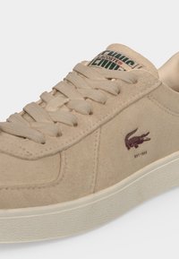 Μπεζ αθλητικό παπούτσι Lacoste με σουέτ υφή, μπεζ κορδόνια, λευκή σόλα και μικρό σκούρο καφέ λογότυπο κροκοδείλου στο πλάι.