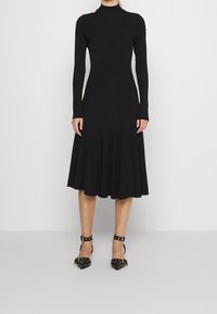 Svart långärmad klänning med en figurnära överdel och utsvängd kjol, tillverkad av strukturerad tyg. Matchas med svarta högklackade skor med nitar.