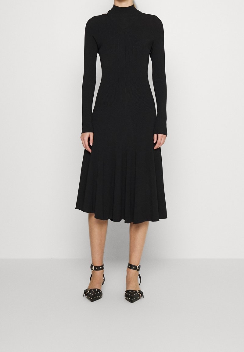 Svart långärmad klänning med en figurnära överdel och utsvängd kjol, tillverkad av strukturerad tyg. Matchas med svarta högklackade skor med nitar.