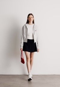 Γκρι φούτερ με φερμουάρ πάνω από λευκό μπλουζάκι, συνδυασμένο με μαύρη φούστα. Λευκά αθλητικά παπούτσια και μια κόκκινη τσάντα ολοκληρώνουν το σύνολο. Ομαλές υφές συνολικά.