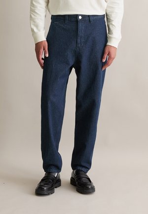Mann trägt dunkelblaue Jeans, weißes langärmeliges Oberteil und schwarze Lederloafers, steht vor einem beigen Hintergrund.