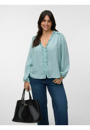 Frau trägt eine hellblaue, rüschige Bluse und blaue Jeans, hält eine große schwarze Handtasche mit Schnallendetail.