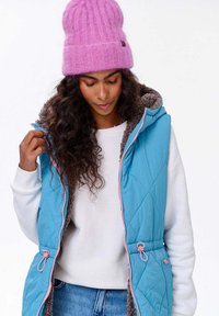 Zateplená vesta v tyrkysové barvě s růžovými akcenty a fleecovou podšívkou, s kapucí a dvěma postranními kapsami. Nosená přes bílou košili.