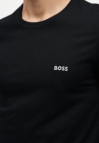 Nahaufnahme einer Person, die ein schwarzes Rundhalsshirt mit dem weiß gestickten Wort „BOSS“ auf der linken Brust trägt.