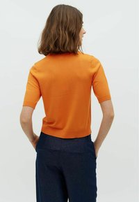 Krátký rukáv oranžový pletený top s kulatým výstřihem, vyrobený z měkkého materiálu, v kombinaci s tmavě modrými kalhotami a jednoduchým, přiléhavým střihem.