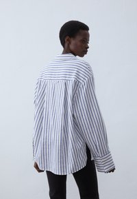 White button-up shirt with blue stripes, featuring long sleeves and a loose fit, with a pointed collar and buttoned cuffs.