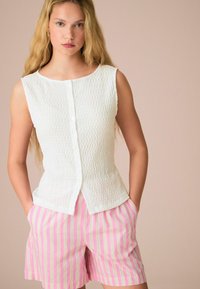 Débardeur texturé blanc sans manches avec des boutons à l'avant, associé à un short rayé rose et blanc avec des poches latérales. Fond clair.