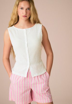 Débardeur texturé blanc sans manches avec des boutons à l'avant, associé à un short rayé rose et blanc avec des poches latérales. Fond clair.
