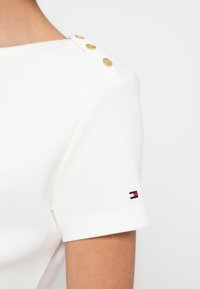 Λευκό βαμβακερό μπλουζάκι με χρυσά κουμπιά στους ώμους και μικρό λογότυπο σε κόκκινο, λευκό και μπλε στο μανίκι. Κοντά μανίκια, εφαρμοστή σχεδίαση.