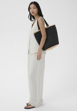 Ázijská žena v bielom prúžkovanom tielku bez rukávov a nohaviciach nesie veľkú čiernu pletenú tašku na ramene.
