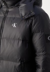 Černá péřová bunda s lesklým povrchem, zipovým zapínáním, odnímatelnou kapucí a logovými detaily na hrudi a rukávu. Obsahuje boční kapsy.