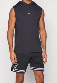 Persona atlética vistiendo una parte superior sin mangas con capucha negra y pantalones cortos de malla negra con detalles de raya blanca, de pie con los brazos relajados.