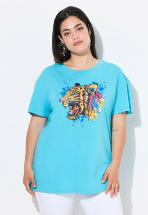 Nainen pukeutunut turkoosinväriseen t-paitaan, jossa on värikäs karjuvan tiikerin kuva ja valkoiset housut, seisoo yksiväristä valkoista taustaa vasten.
