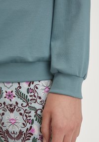 Lichtblauwe sweatshirt met een zachte textuur en aansluitende boorden, gecombineerd met een lichtblauwe stof met een patroon van bloemen en paarse en groene accenten.