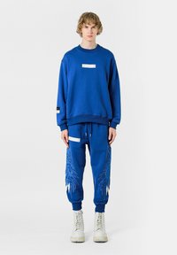 Sudadera azul y joggers a juego con bordados que parecen plumas. Presenta un parche en la sudadera y una etiqueta decorativa en los pantalones.