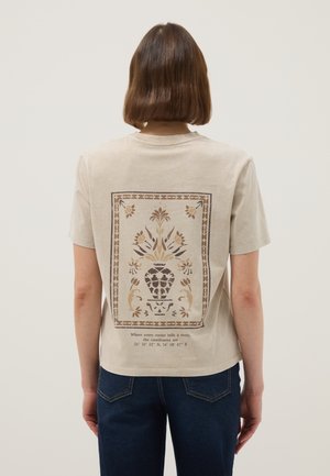 Persoon met kort bruin haar die een beige T-shirt draagt met een decoratieve bruine vaas met bloemenontwerp en coördinaten op de achterkant, gecombineerd met een donkere spijkerbroek.