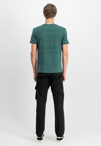 Zelené bavlněné tričko s texturovaným potiskem na zádech a grafikou inspirovanou armádou; spárováno s černými cargo kalhotami a bílými teniskami.