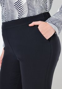 Marina blå byxor i en slät, elastisk tygkvalitet, med en platt midja och en sidficka. Kombinerat med en mönstrad blus.