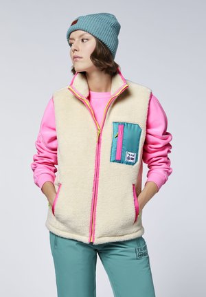 Young woman wearing a teal beanie, pink sweatshirt, cream fleece vest with teal and pink pocket, and teal pants, standing with hands in pockets.