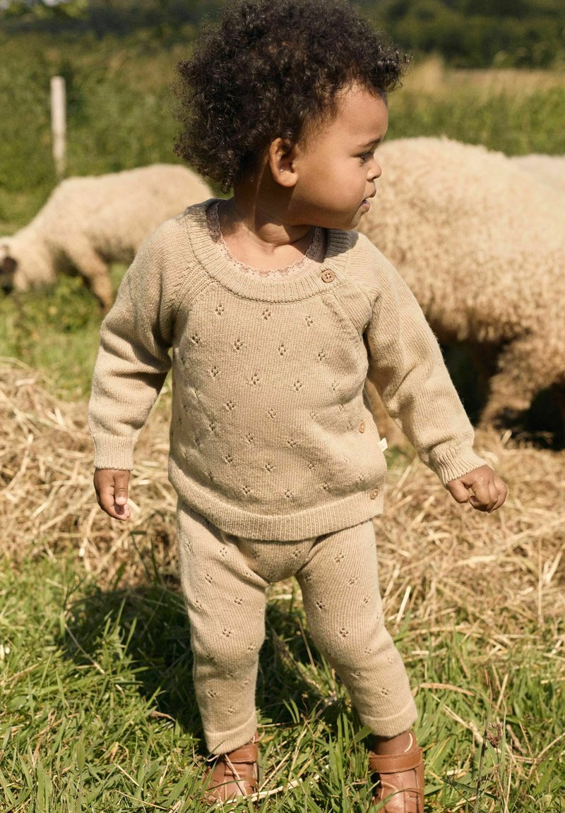 Un bambino in una tuta di maglia beige e scarpe marroni sta in piedi sull'erba con delle pecore che pascolano dietro di lui in un ambiente esterno soleggiato.
