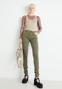 Top sin mangas gráfico beige superpuesto sobre una camisa de manga larga marrón, jeans skinny verde oliva y zapatos de plataforma negros. Sostiene un bolso beige claro.