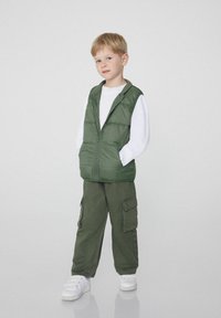 Zelená vycpaná vesta se zipem nošená přes bílou halenku s dlouhým rukávem, doplněná o tmavě zelené cargo kalhoty a bílé tenisky.