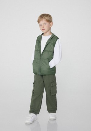 Grüne gepolsterte Weste mit Reißverschluss, getragen über einem weißen Langarmshirt, kombiniert mit dunkelgrünen Cargo-Hosen und weißen Sneakers.