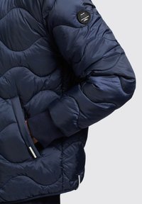 Mörkblå puffjacka med ett kviltat mönster, som har en logopatch, dragkedjefickor och ribbade ärmslut för extra värme.