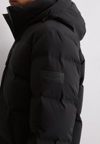 Schwarze Daunenjacke mit matter Oberfläche, ausgestattet mit einem hohen Kragen, verstellbare Kapuze und einem Logo-Patch am linken Ärmel.