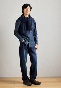 Ναυτική πουκάμισο με κουμπιά και μικρά λουλουδάτα σχέδια, συνδυασμένο με σκούρα μπλε φαρδιά τζιν και πλεκτό ναυτικό φουλάρι ριγμένο στους ώμους.