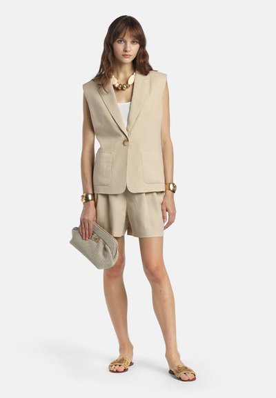 Kvinde iført beige ærmeløs blazer, matchende shorts, hvid top, stor skalle halskæde, guldarmbånd, holder vævet clutch, stående i sandaler.