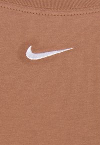 Brązowa materiał o prążkowanej teksturze z drobną fakturą, z białym haftowanym logo Nike umieszczonym po lewej stronie.