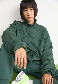 Πράσινο ζακέτα με φερμουάρ και υφή, ταιριαστά παντελόνια και χέρι που ακουμπάει στο γόνατο. Τα παπούτσια είναι λευκά με έντονα πράσινα στοιχεία.