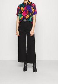 Camicia a maniche corte multicolore con motivo floreale su sfondo nero, abbinata a pantaloni neri a vita alta con gamba larga e stivaletti neri.