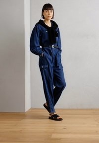 Denim jumpsuit med lange ermer og innsnevret midje, med lommer, iført svart skjerf og svarte sandaler, i en minimalistisk setting.