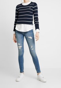 Granatowy sweter w paski z białymi i różowymi liniami, noszony na białej koszuli z długim rękawem. Do tego jasnoniebieskie podarte dżinsy i białe trampki.