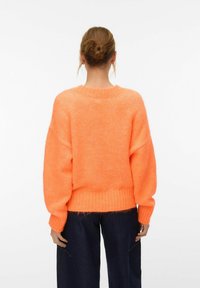 Femme aux cheveux attachés en chignon, portant un pull orange vif en tissu duveteux et un pantalon bleu foncé, tournée de dos devant un fond blanc uni.