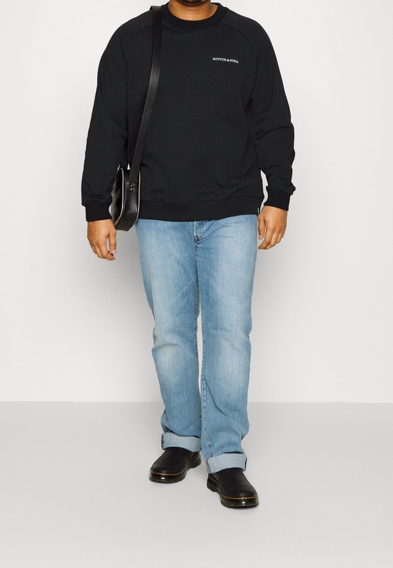 Felpa nera con polsini a coste, logo grafico blu, jeans azzurri e scarpe nere. Una borsa a tracolla pende dalla spalla sinistra.