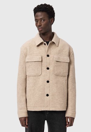 Beige uldblandet jakke med krave, to brystlommer, sorte knapper og lige snit. Båret over en mørk skjorte og jeans.