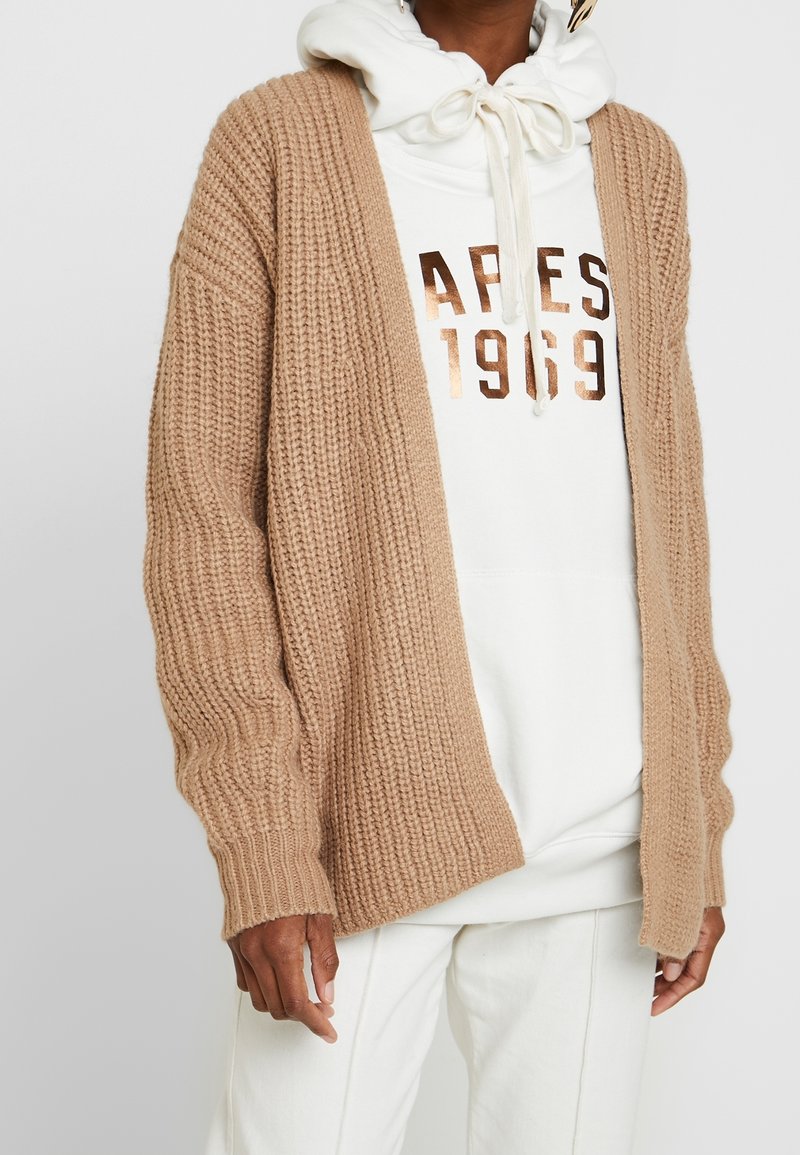 Personne portant un cardigan en maille épaisse beige par-dessus un sweat à capuche blanc avec l'inscription "APES 1969" en lettres métalliques, assorti d'un pantalon blanc.
