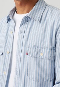 Camicia azzurra chiara con righe verticali bianche, tessuto morbido, maniche lunghe e una sola tasca sul petto con etichetta del marchio.