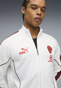 Hvid lynlåsjakke med sorte detaljer og røde logoer, herunder "KING" og et løvedesign. Stoffet ser glat ud med en atletisk pasform.